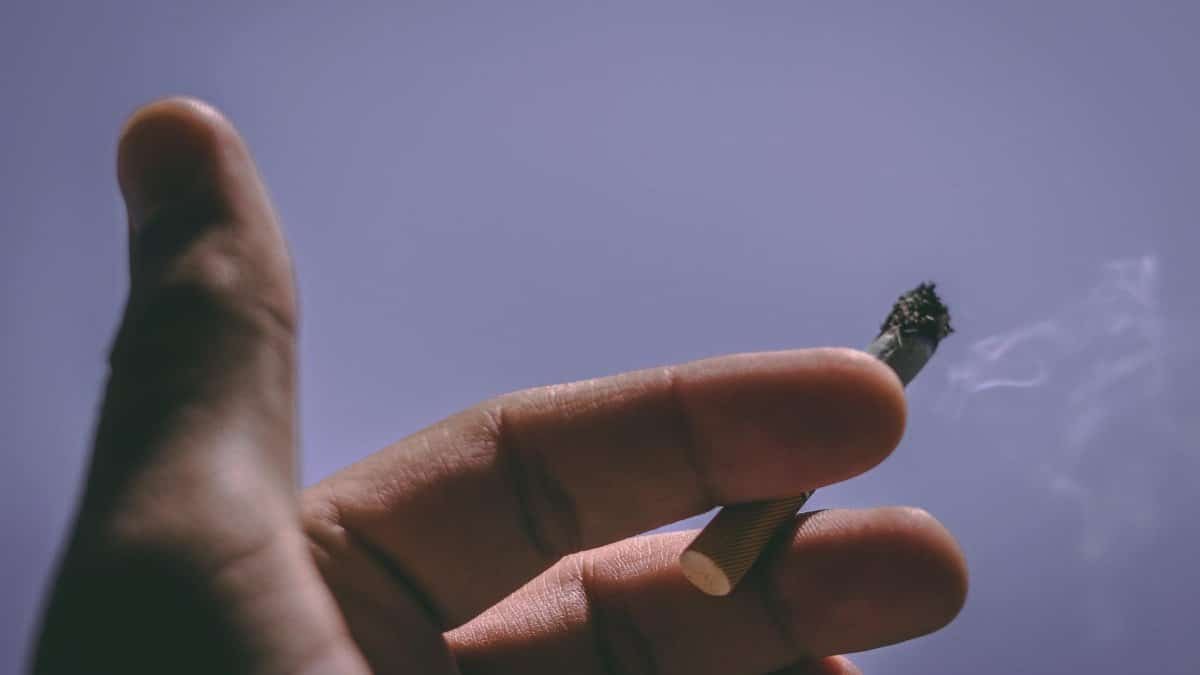 Close-up of a hand holding a lit cigarette against a clear blue sky.