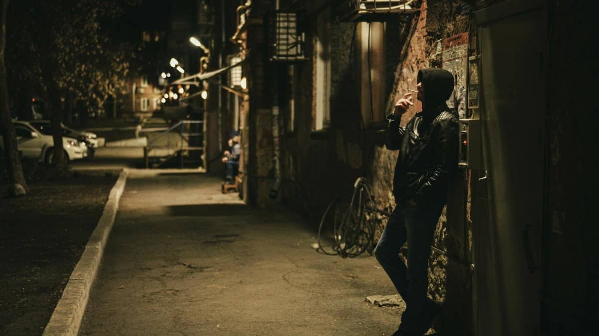A solitary figure in a hoodie smokes at night in a dimly lit alley.