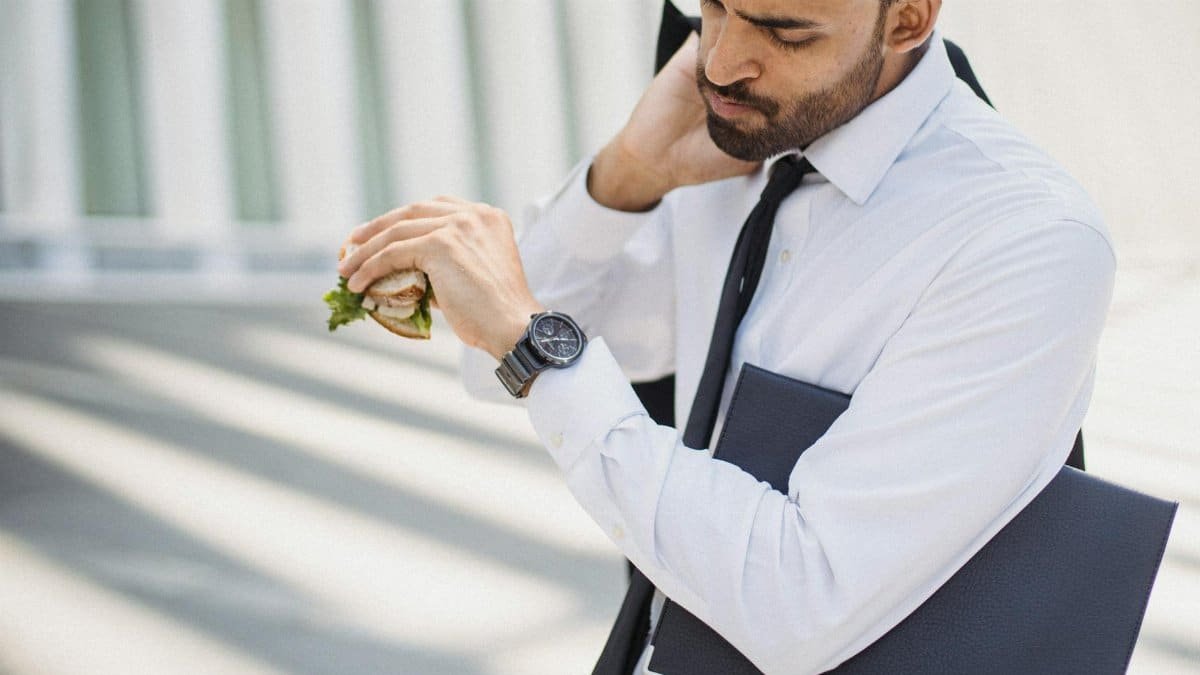 Professional man in corporate attire checking time while holding a sandwich outdoors.
