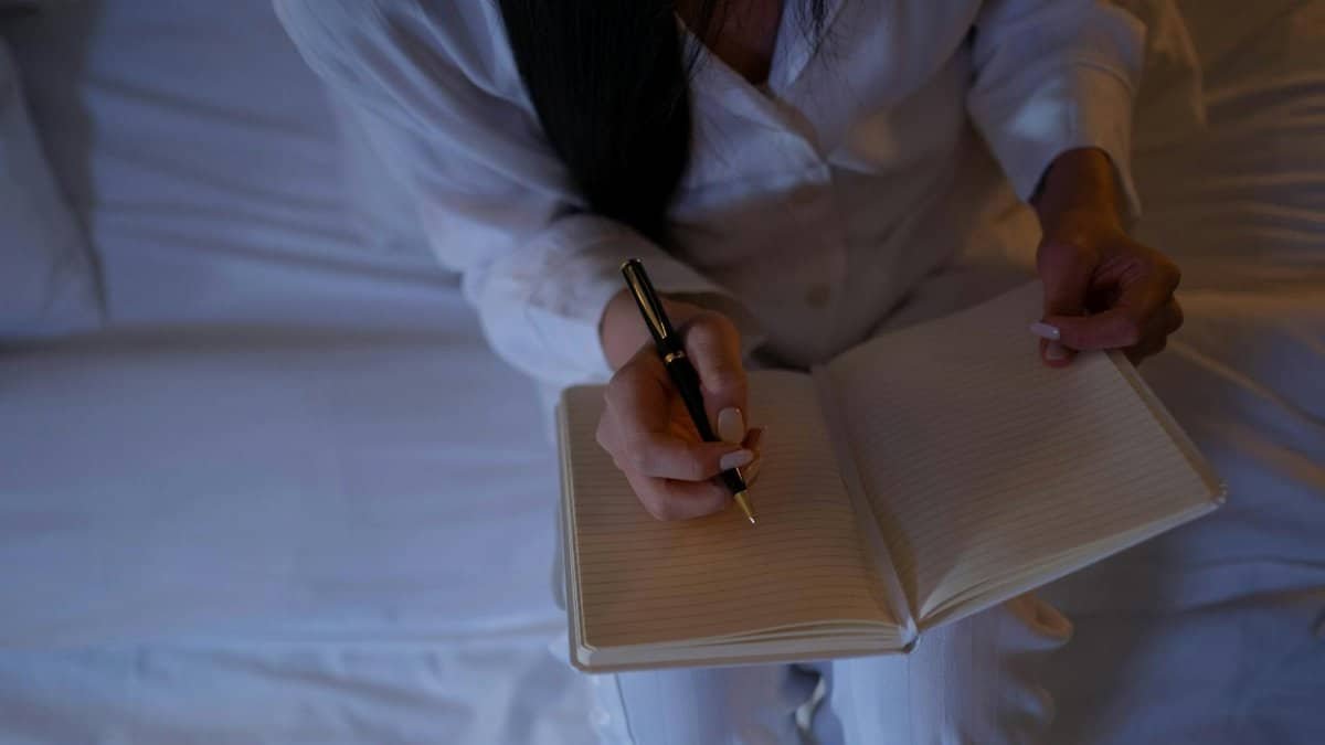 A person is writing in a notebook while sitting on a bed in a dimly lit room.