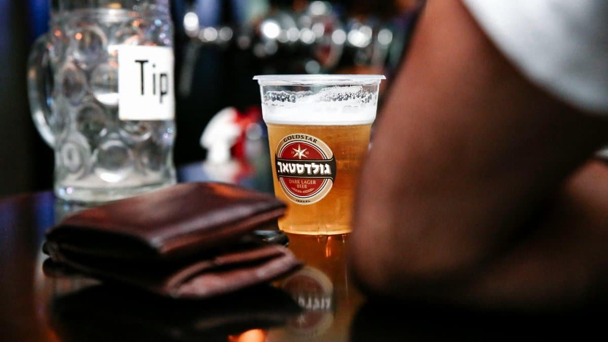 A close-up shot of a beer in a plastic cup on a bar counter, with a tip jar and wallet nearby.