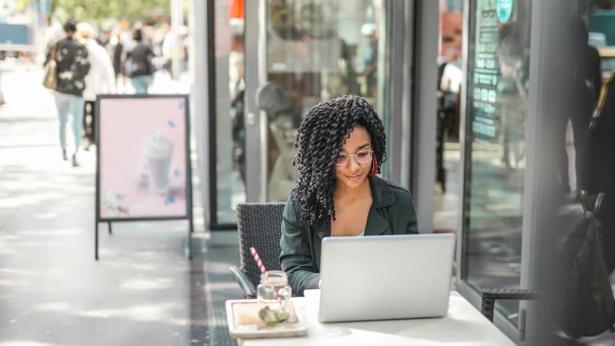 High angle of pensive African American female freelancer in glasses and casual clothes focusing on screen and interacting with netbook while sitting at table with glass of yummy drink on cafe terrace in sunny day