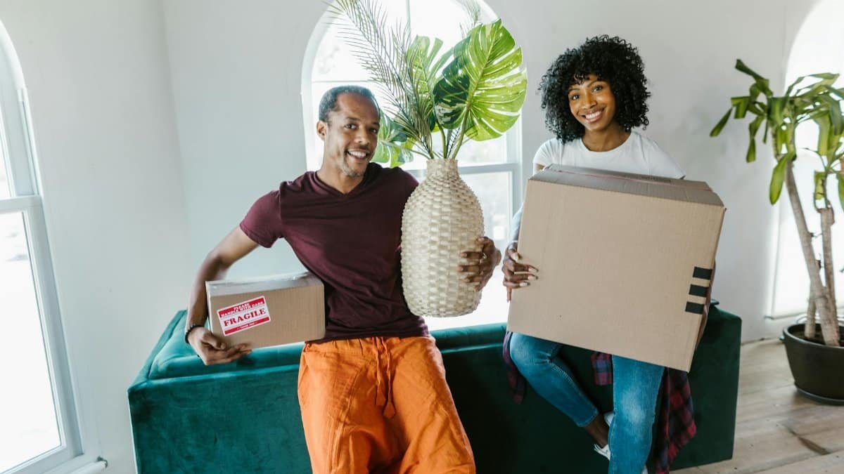 Couple smiling while moving into a new home, carrying boxes and a plant.