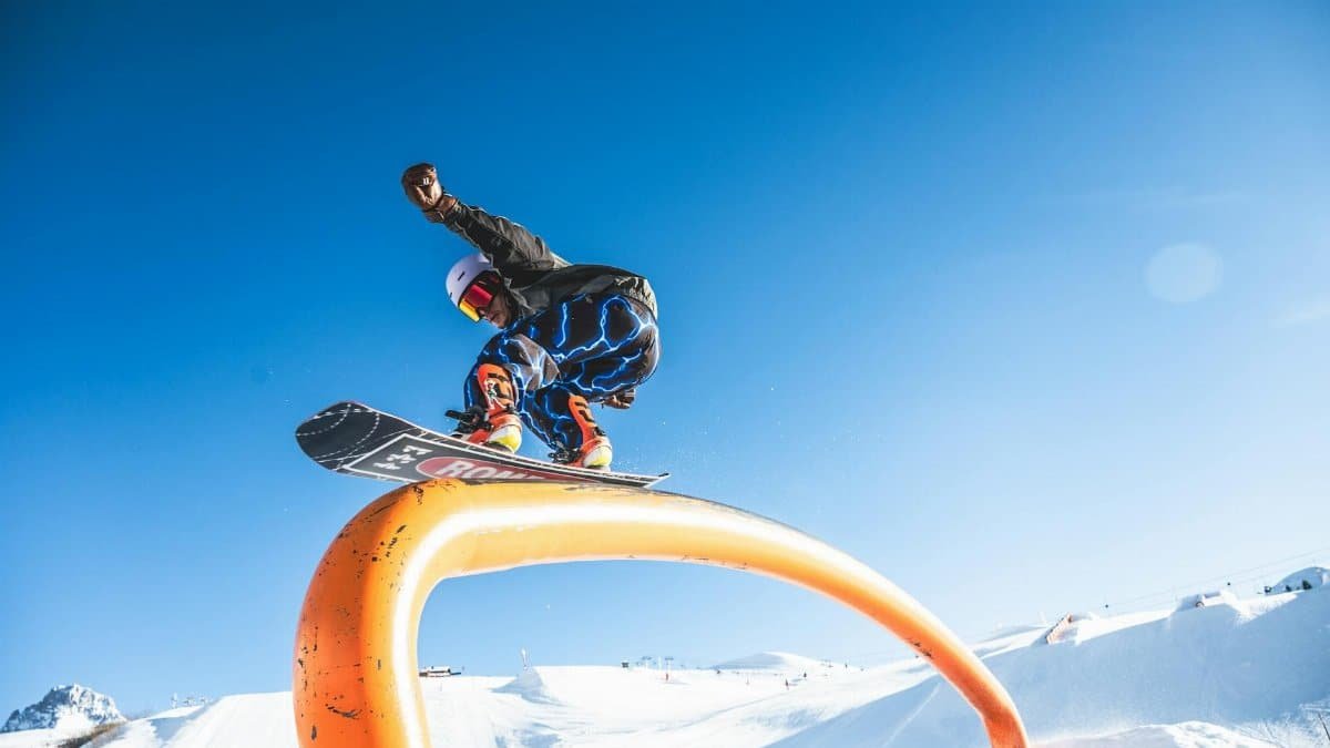 Snowboarder performing a trick over a rail in a snowy landscape with blue skies.
