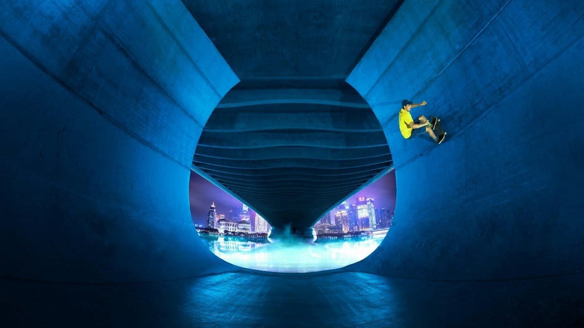 A skateboarder performs an extreme trick under a bridge with a city skyline backdrop, capturing urban adventure.