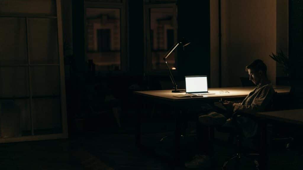 A man working late at night in a dimly lit office, with a bright laptop screen.