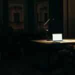 A man working late at night in a dimly lit office, with a bright laptop screen.