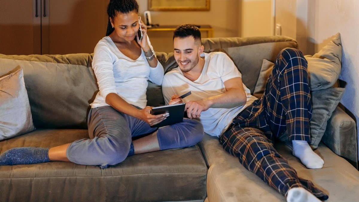 Interracial couple laughing and shopping online at home using tablet and smartphone.