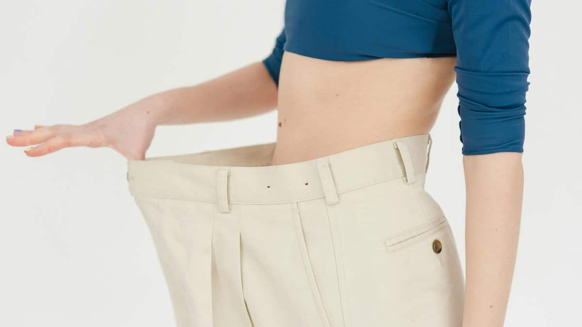 A woman demonstrates significant weight loss by wearing oversized pants indoors.