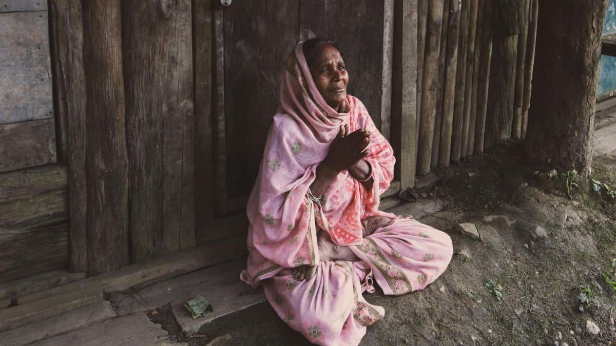 Elderly woman sitting outdoors in prayer by a wooden doorway, signifying resilience and hope.