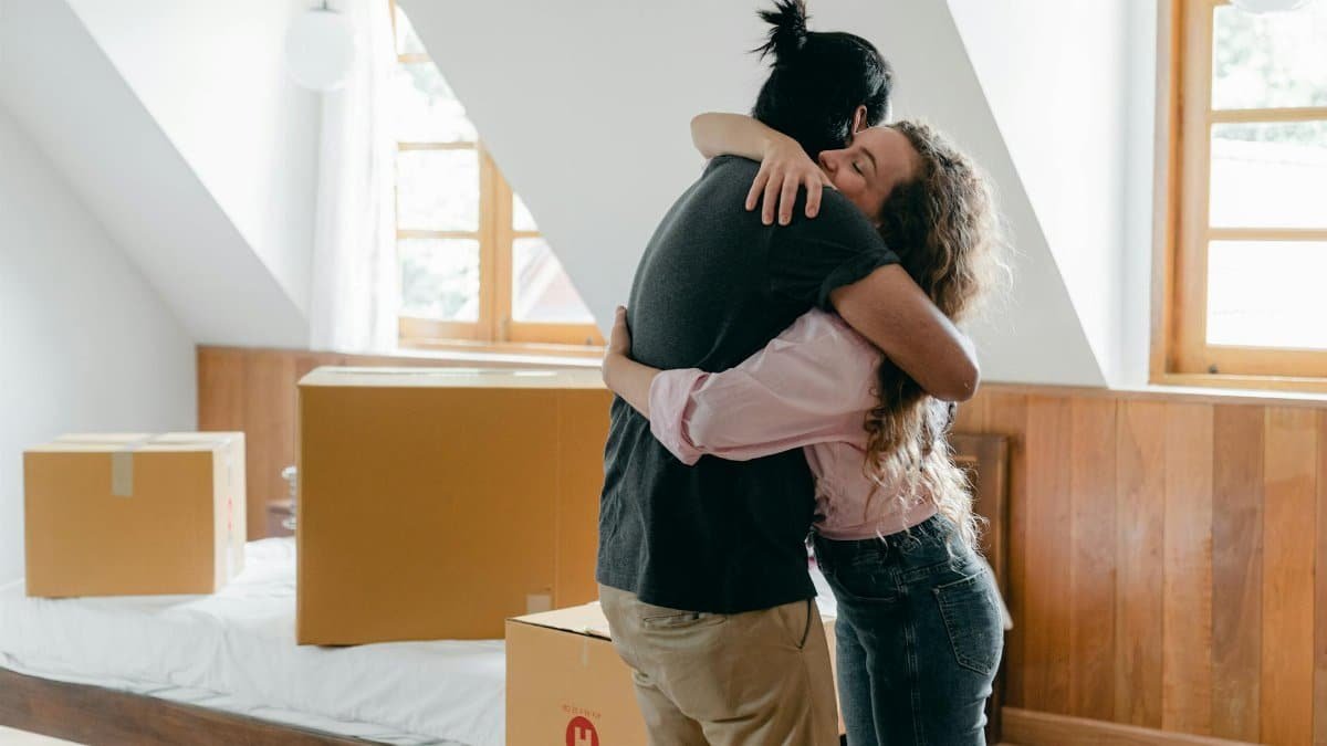 Side view of young multiethnic couple embracing each other while finishing relocation and standing near carton boxes in light spacious bedroom