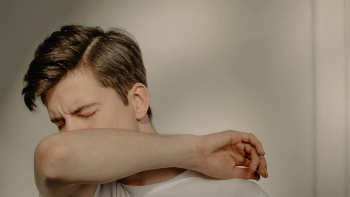 A young man sneezes into his elbow, showcasing a hygiene practice, in an indoor setting.
