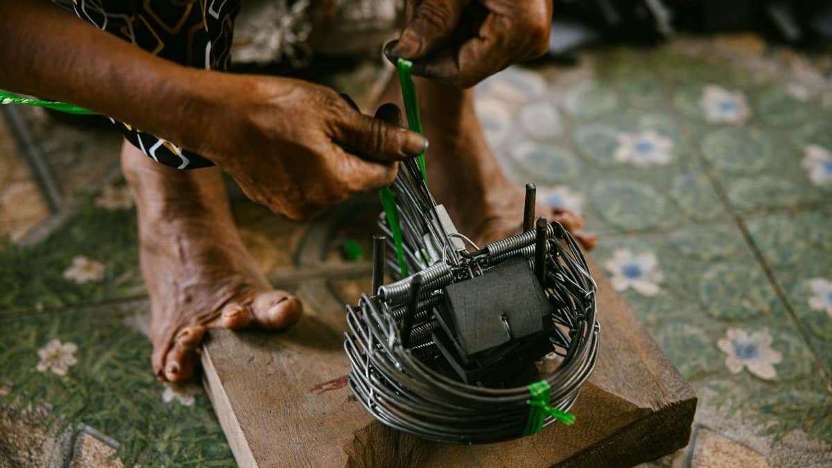 Craftsman's hands skillfully weaving a metal trap on wooden base indoors. Traditional craftsmanship.