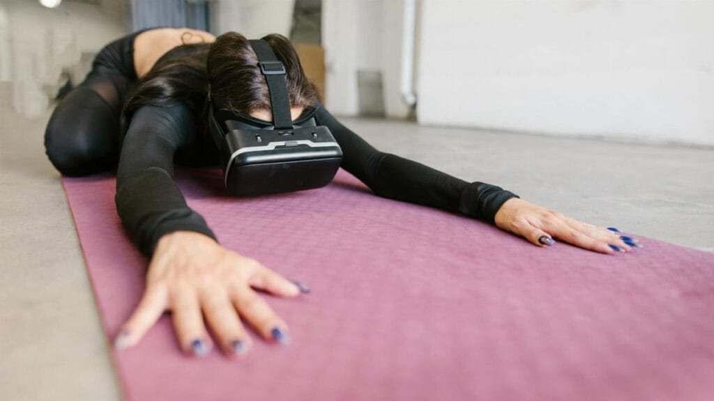 Adult woman in yoga pose using VR headset on pink mat, exploring virtual reality fitness.