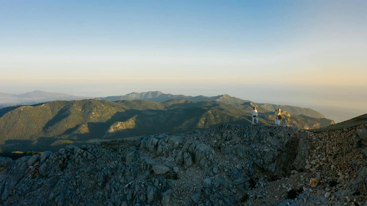 Two hikers reach a rocky mountain summit under a clear sky at sunset.