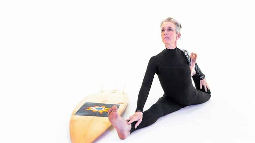 Senior woman in wetsuit practicing yoga stretches beside a surfboard on a white background.