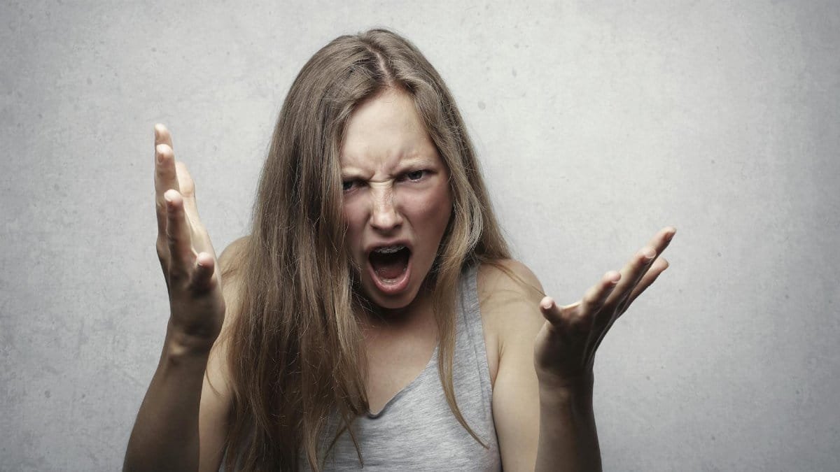 A young woman showing intense anger and frustration, yelling with hands raised against a gray background.