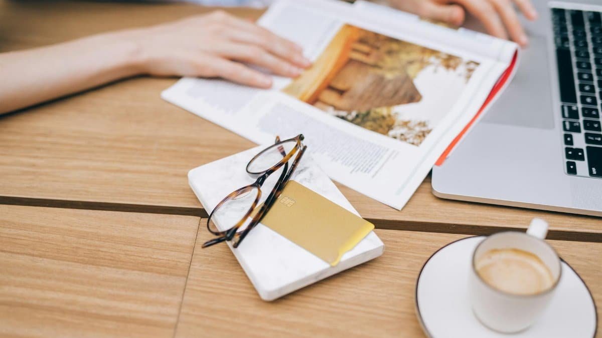 A warm workspace featuring a laptop, coffee cup, magazine, and eyeglasses on a wooden table.