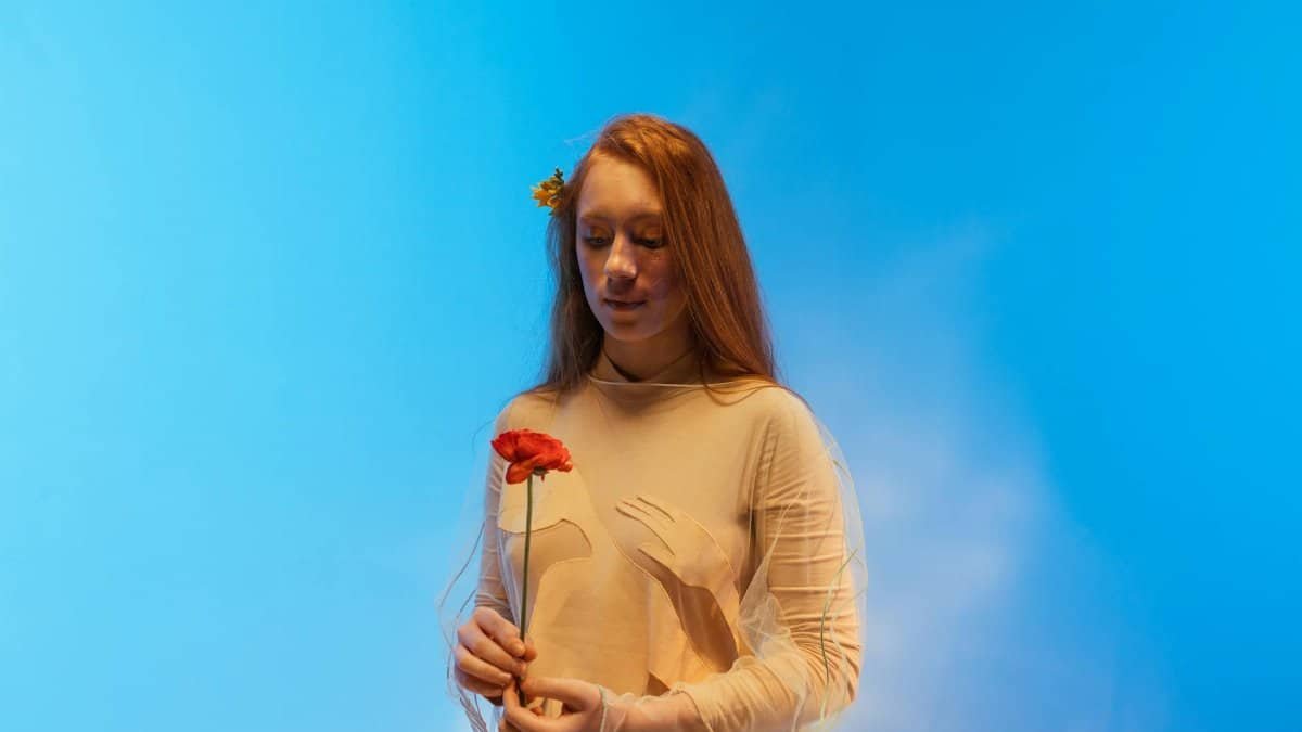 A young woman in a beige dress holds a red flower against a vibrant blue sky background, embodying a serene and conceptual art style.