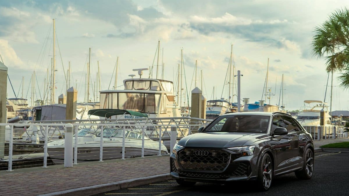 Sleek luxury car parked beside yachts in a scenic marina setting under a cloudy sky.