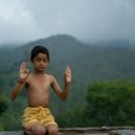 A young boy practices meditation outdoors with a peaceful mountain backdrop.