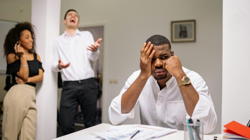 A man at a desk looks frustrated while two coworkers laugh. Office workplace conflict concept.