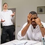 A man at a desk looks frustrated while two coworkers laugh. Office workplace conflict concept.