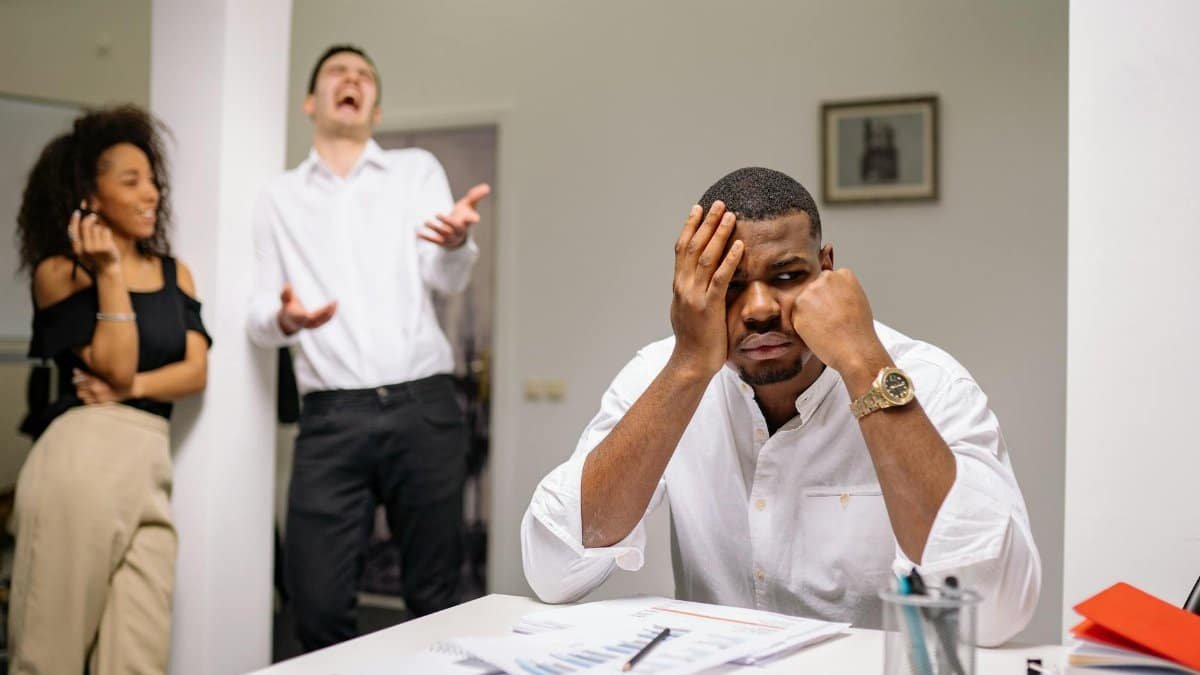 A man at a desk looks frustrated while two coworkers laugh. Office workplace conflict concept.
