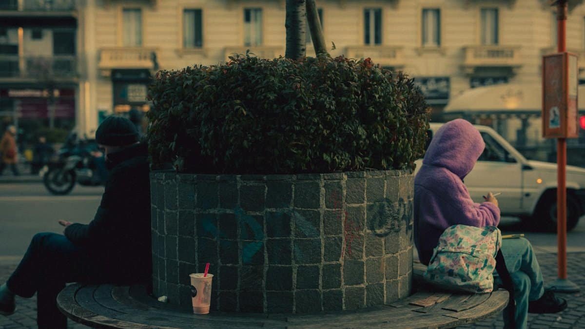 A city street scene in Milan showing two people seated on opposite sides of a planter.