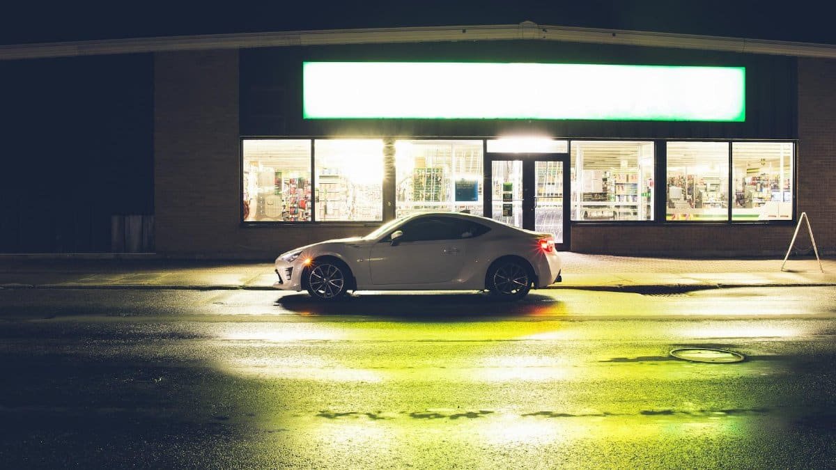 White coupe car on roadside at glowing shop with signboard late at night time