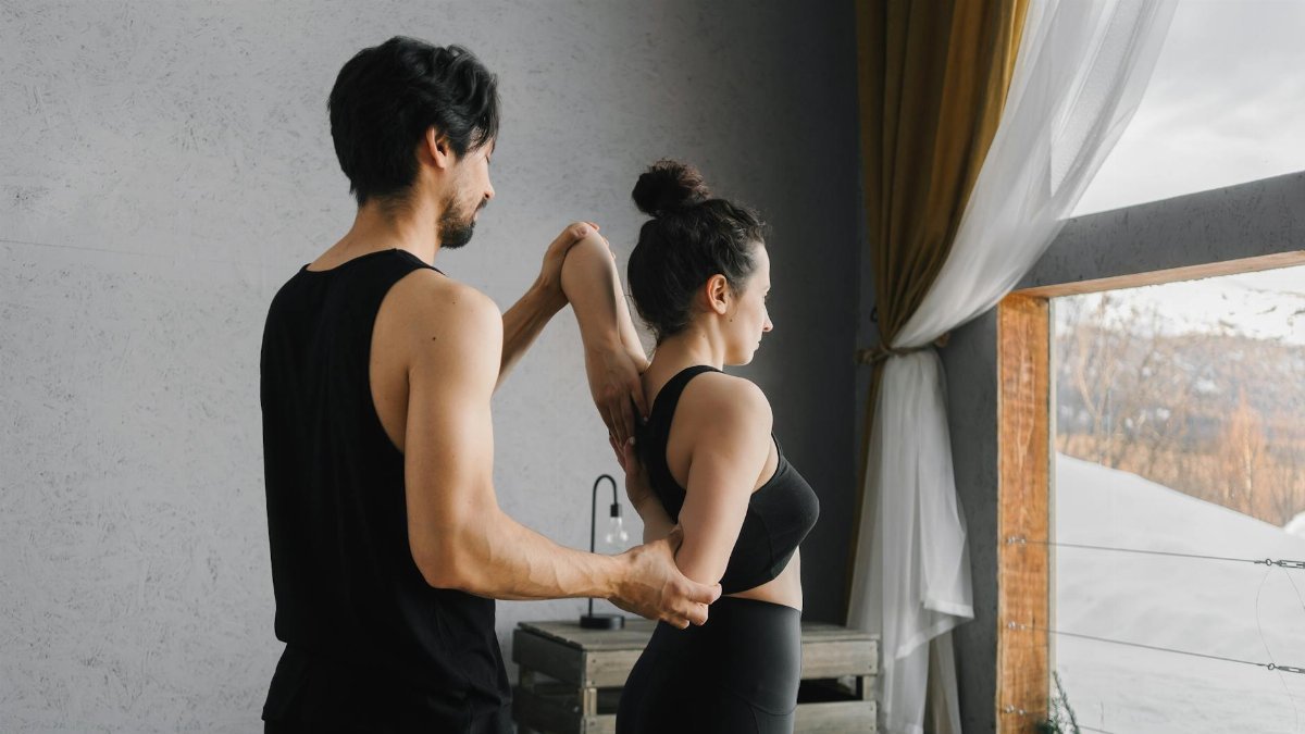 A man and woman practicing partner yoga poses near a large window inside a cozy room.