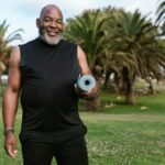Smiling elderly man holding a yoga mat in a sunny park surrounded by palm trees.