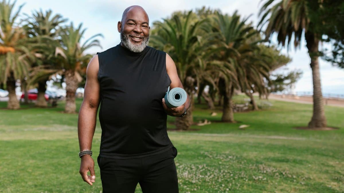 Smiling elderly man holding a yoga mat in a sunny park surrounded by palm trees.