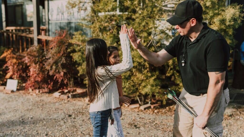 An instructor high-fiving a child at an archery range during a sunny day.