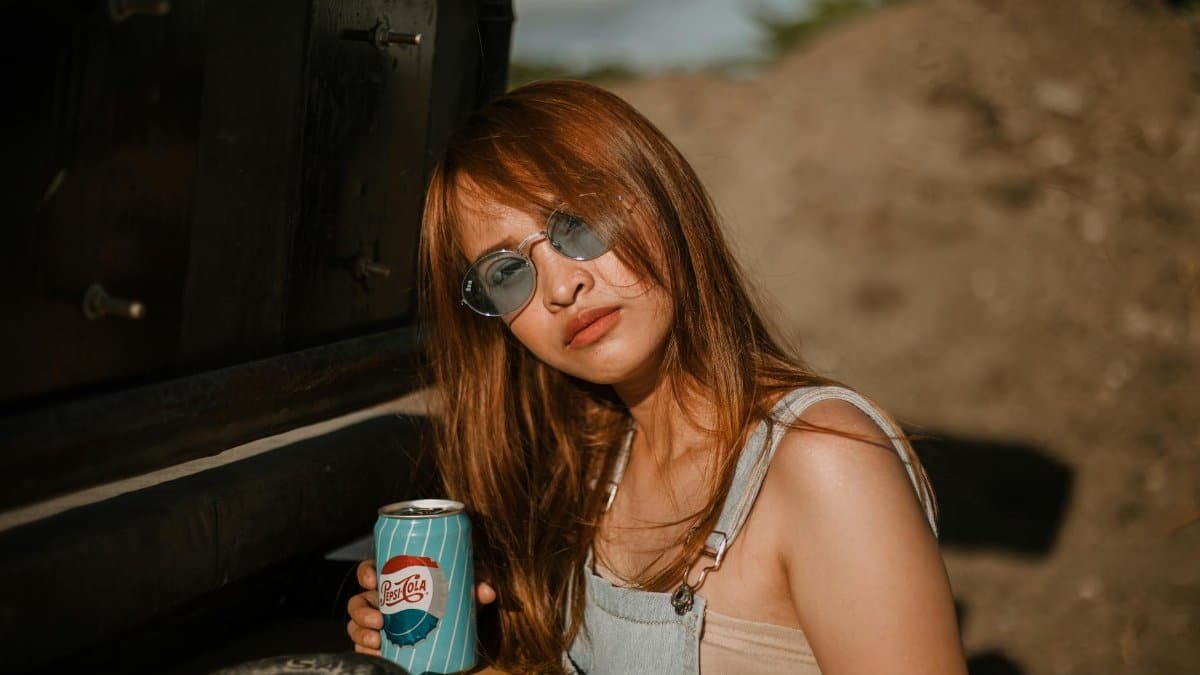Young woman with red hair and sunglasses holding a soda can outdoors in sunlight.