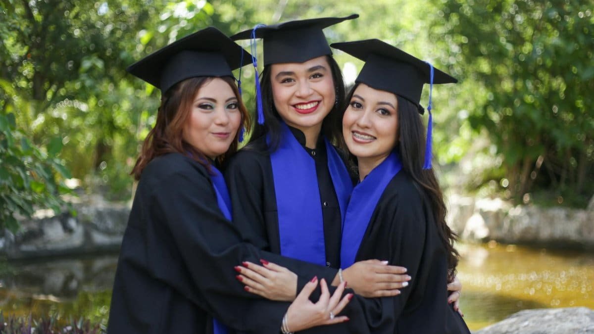 Three cheerful female graduates in caps and gowns embracing with bright smiles in an outdoor setting.