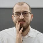 A man with eyeglasses and beard shows discomfort from a toothache indoors.