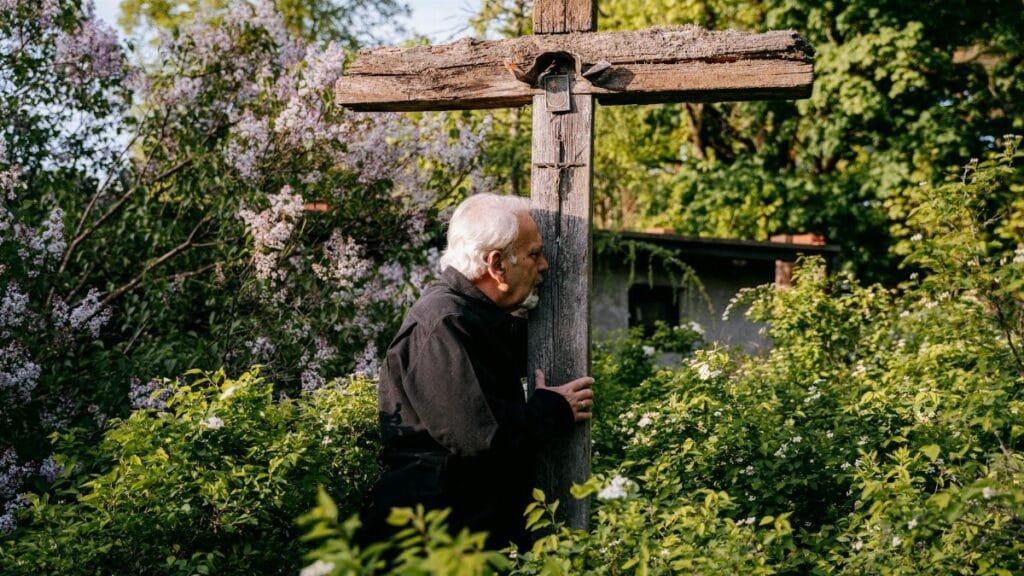 Elderly man holds a wooden cross in a lush outdoor garden, depicting reflection and grief.