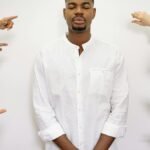 A calm man in white stands with closed eyes while several hands point at him in concept of accusation.