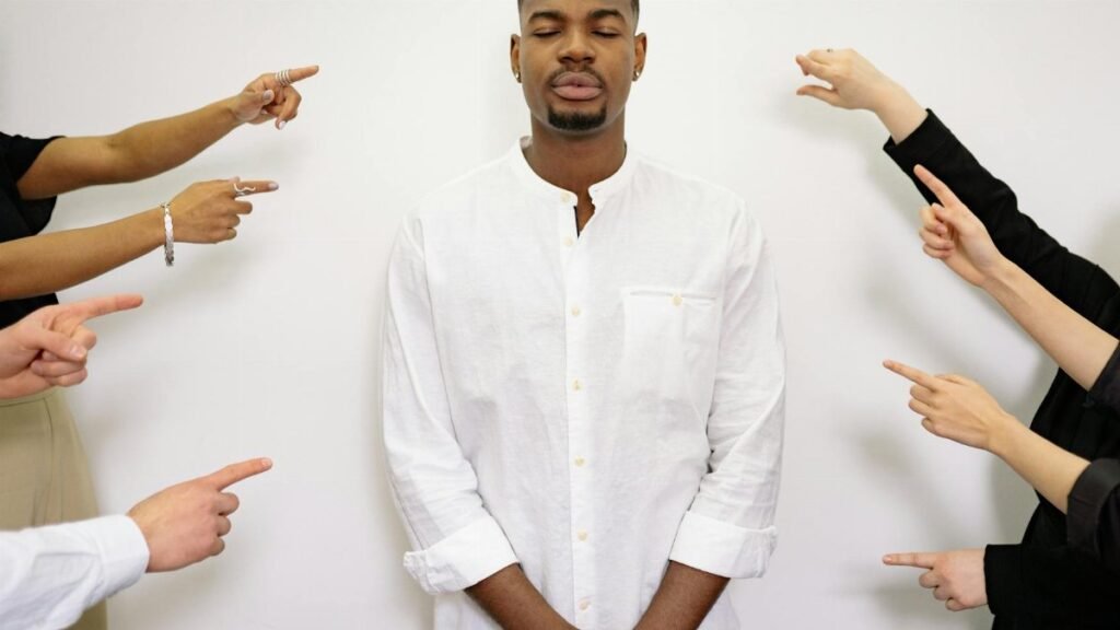 A calm man in white stands with closed eyes while several hands point at him in concept of accusation.