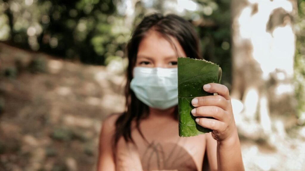 A young girl wearing a face mask holds a green leaf in a forest setting, reflecting nature and new health norms.