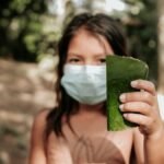A young girl wearing a face mask holds a green leaf in a forest setting, reflecting nature and new health norms.