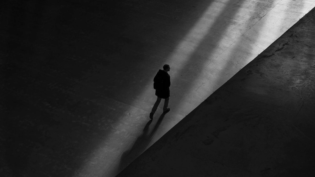 Black and white photo of a man walking through shadows on a deserted pavement, creating a moody silhouette.