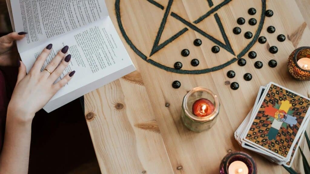From above of crop unrecognizable fortune teller reading textbook at table with tarot cards and candles during divination process