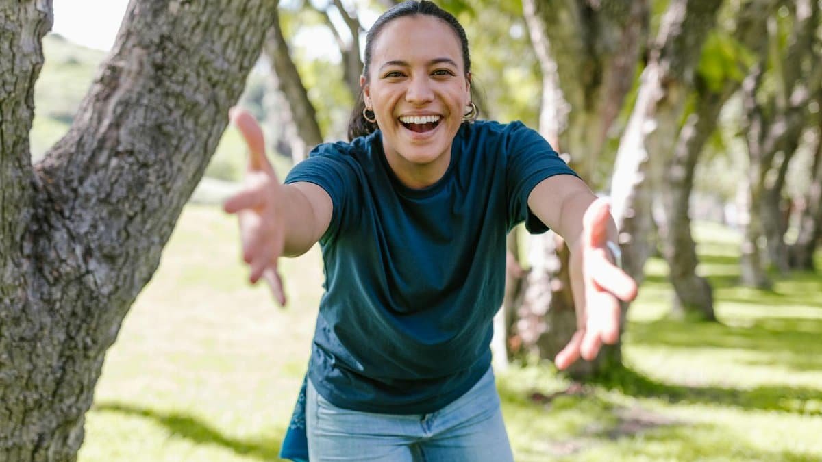 Smiling woman reaching out with open arms in a sunny, green park setting.