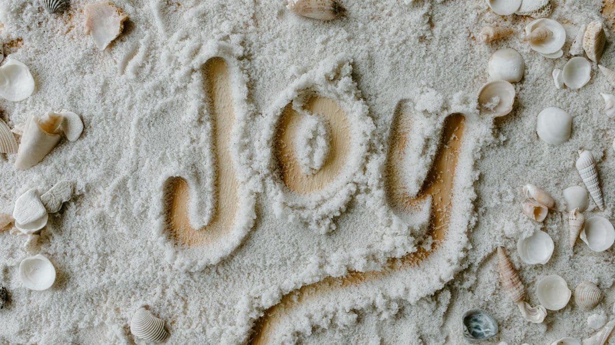 The word 'joy' written in sand with various seashells scattered around.