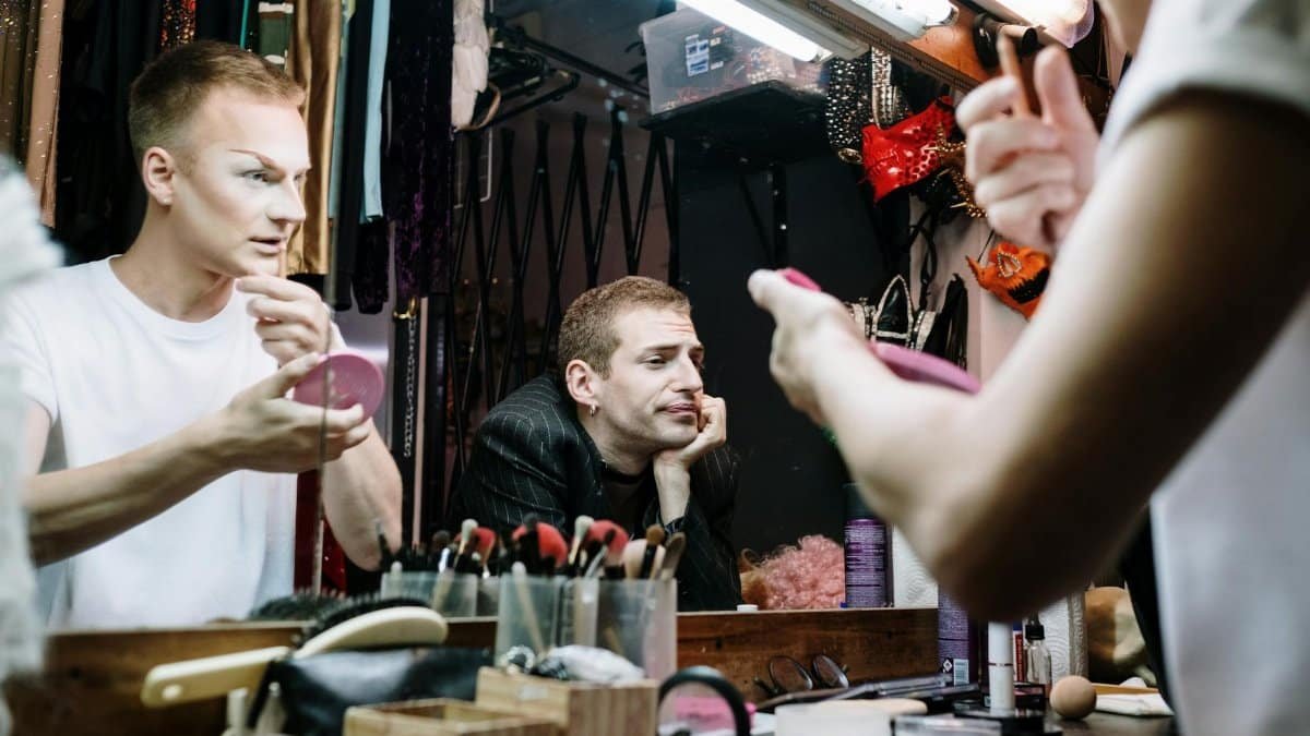 Two drag performers apply makeup backstage, showcasing identity and transformation.