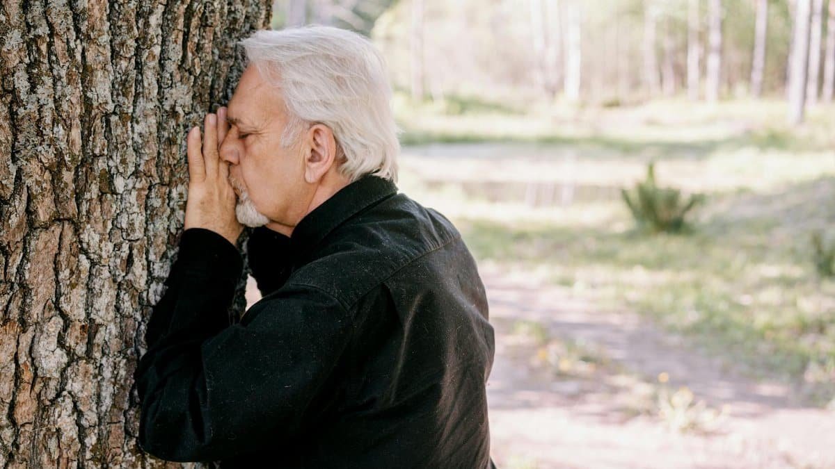 Senior man in black shirt leans on tree trunk, expressing contemplation outdoors.