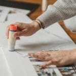Crop anonymous person in warm gray wear applying glue stick on papers and arranging photo album on floor