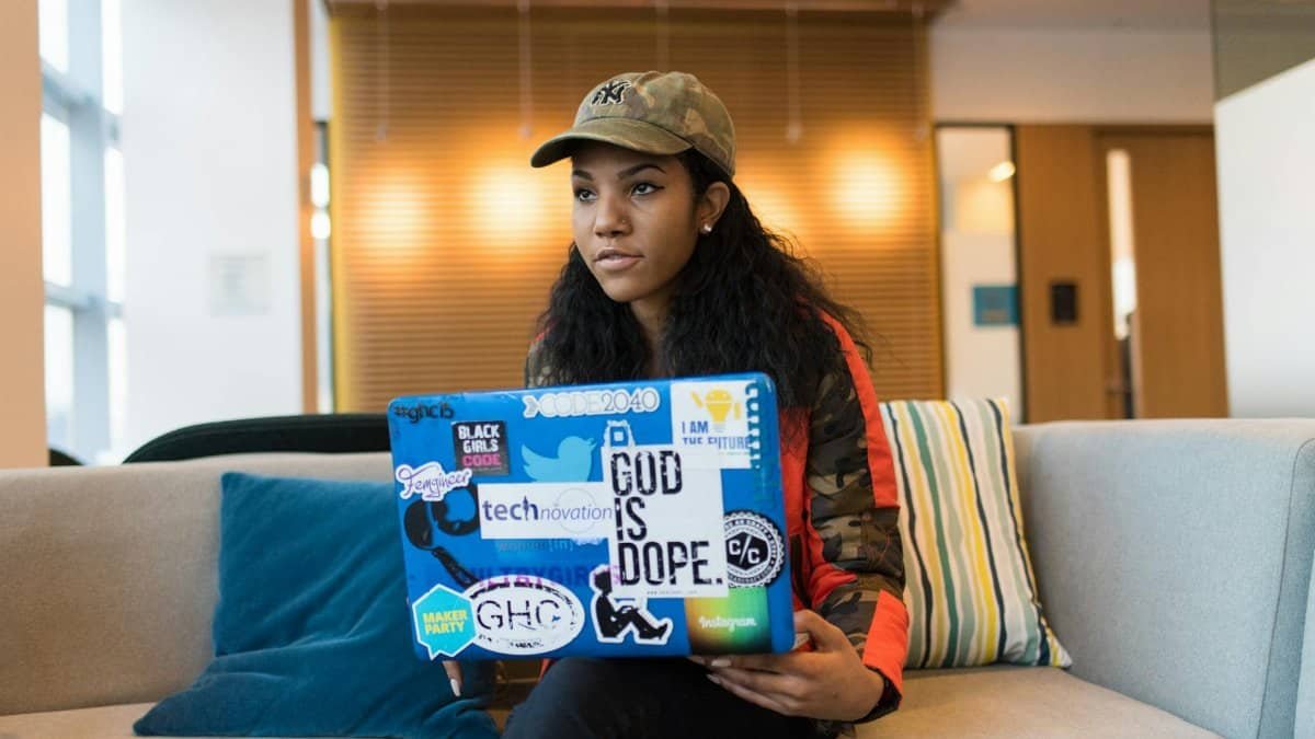 Young woman in camo jacket and cap using a sticker-covered laptop indoors, focused on work.
